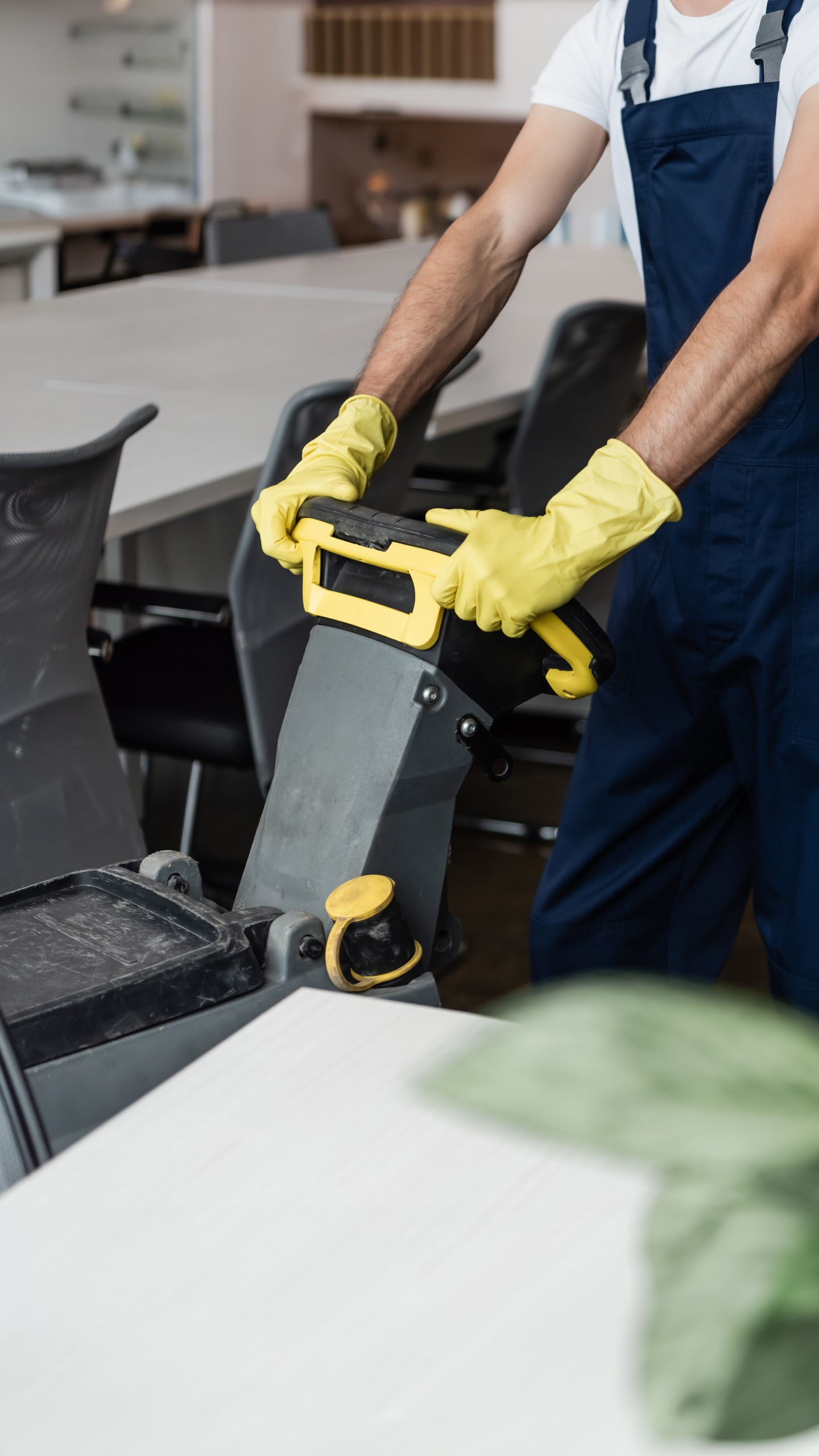 A person wearing yellow gloves is operating a floor cleaning machine in a bright, modern workspace.